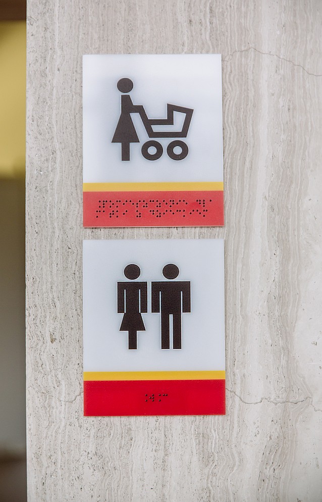 Na ścianie wiszą dwie ikonki przedstawiające kobietę z wózkiem dziecięcym i toalety. Tabliczki są opisane w brajlu