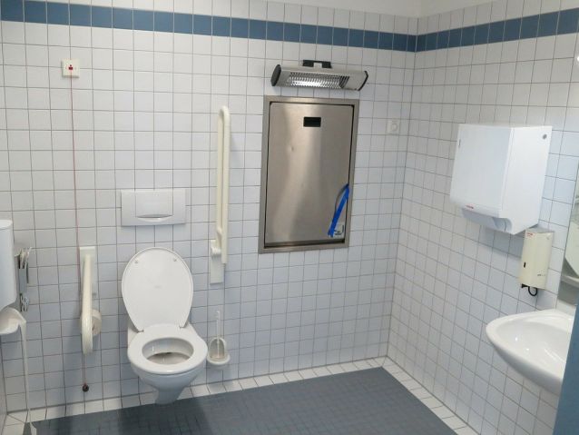 Wnętrze dostosowanej toalety. Na wprost muszla ustępowa z poręczami, po prawej umywalka z poręczami. Obok muszli wisi czerwona linka alarmowa