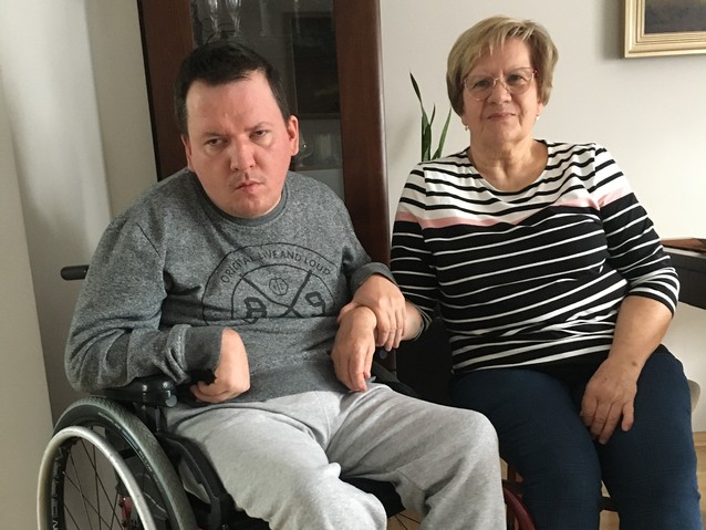 Barbara Bigoszewska siedzi na krześle w pokoju obok na wózku jej syn Bartosz, trzyma ją za rękę