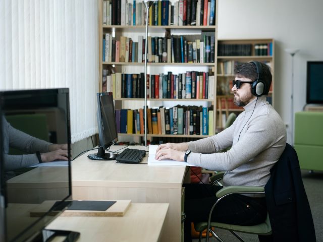 mężczyzna w ciemnych okularach siedzi i czyta brajlem przed nim komputer obok drugie stanowisko w tle półki z kiążkami