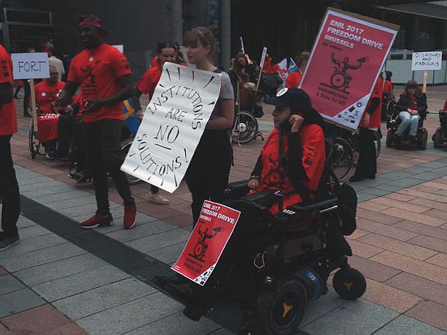 Manifestacja osób z niepełnosprawnością w Brukseli. Na pierwszym planie kobieta na wózku, za nią stoi kobieta z transparentem z napisem po angielsku: instytucje nie są rozwiązaniem
