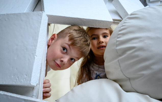 dziewczynka i chłopiec wyglądają zza bialych kartonów i poduszek