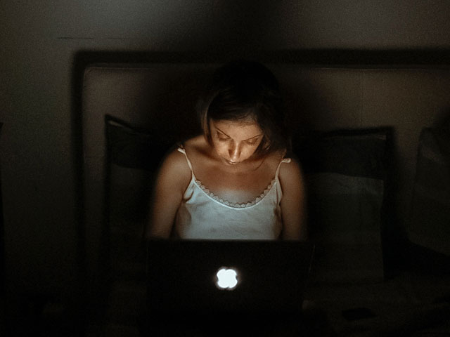 kobieta siedzi na łóżku w ciemności, pisze na komputerze, jest zmęczona