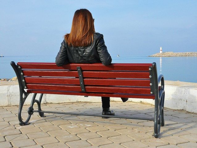 Kobieta siedzi na ławce na brzegu morza. W oddali latarnia morska
