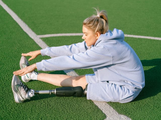 dziewczyna z protezą siedzi na boisku i wyciąga ręce do nóg, ćwiczy