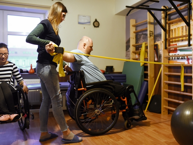 sala rehabilitacyjna kobieta i mężczyzna na wózku mężczyzna wykonuje ćwiczenia z taśmą przy drabinkach przy pomocy rehabilitantki