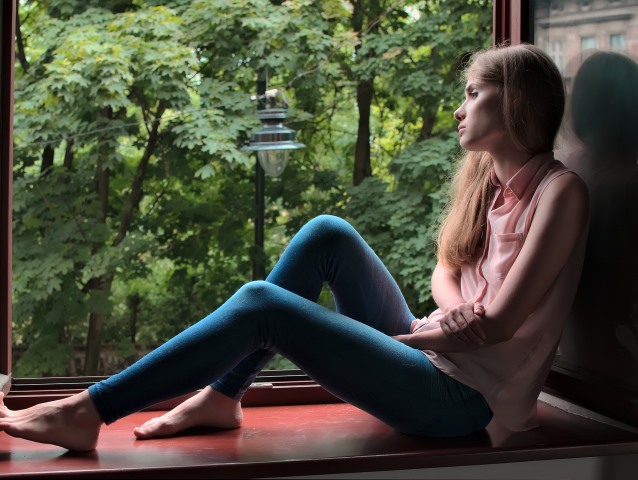 zamyślona młoda kobieta siedzi na parapecie w otwartym oknie, za nim zieleń
