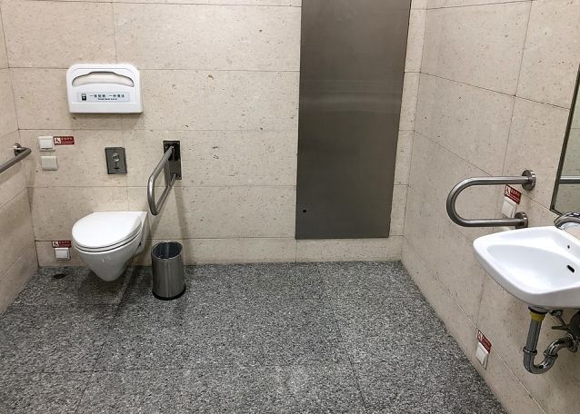 Wnętrze dostosowanej toalety z muszlą ustępową, umywalką i poręczami. W czterech miejscach na ścianie są przyciski alarmowe