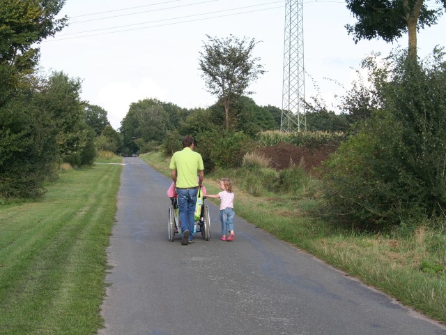 mężczyzna pcha wózek inwalidzki obok idzie mała dziewczynka widoczni od tyłu wokół zieleń