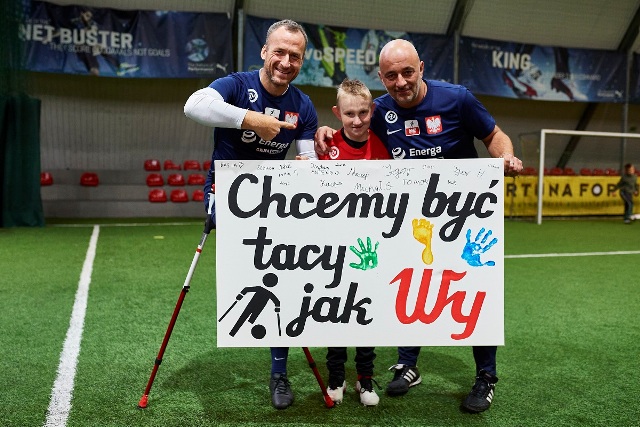 Trzech ampfutbolistów. W środku chłopiec, a po bokach dojrzali mężczyźni. Uśmiechają się szeroko. Trzymają tablicę z napisem: chcemy być tacy jak Wy 