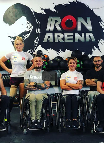 Zdjęcie. Siłownia. Rząd czterech osób pod ścianą z napisem Iron Arena. Trzy osoby są na wózkach, kobieta stojąca ma protezę nogi. Ubrani w sportową odzież
