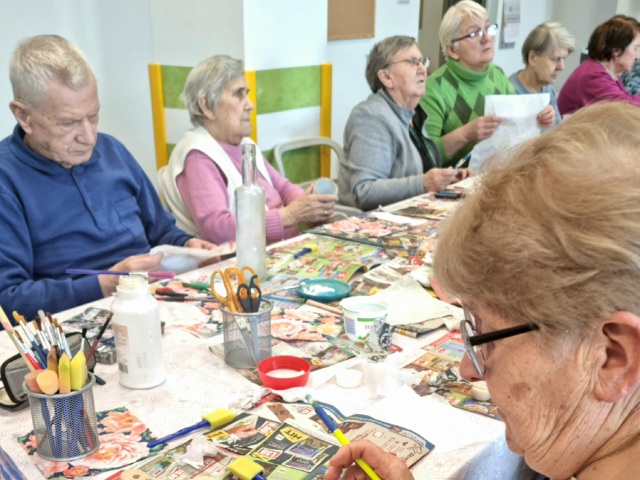 Osoby starsze siedzące przy stole podczas prac artystycznych. Na stole leżą przybory artystyczne,