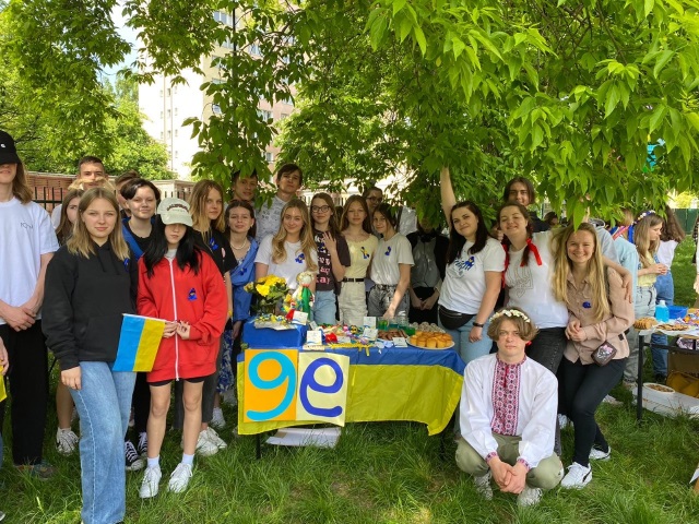 Uczniowie Ukraińskiej Szkoły w Warszawie. Stoją przy ławce pod drzewem. Ławka jest przykryta obrusem w kolorach ukraińskiej flagi. Na ławce są talerze ze słodyczami.