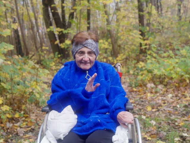 Starsza kobieta siedząca na wózku. Ma krótkir, brązowe włosy i chustke na głowie. Ma na sobie gruby, niebieski polar. 