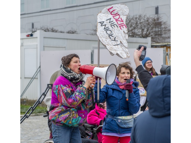 Dwie organizatorki protestu: Joanna Dryjańska i Karolina Kosecka stoją pod Pałacem Prezydenckim. Joanna trzyma transparent w kształcie skrzydeł z napisem "Ludzie nie anioły" a Karolina krzyczy przez megafon. 