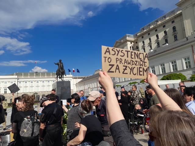 Manifestujący stoją przed Pałecem Prezydenckim. Jedna z osób trzyma podniesioną w górę kartke z napisem "Asystencja to prawdziwe za życiem"