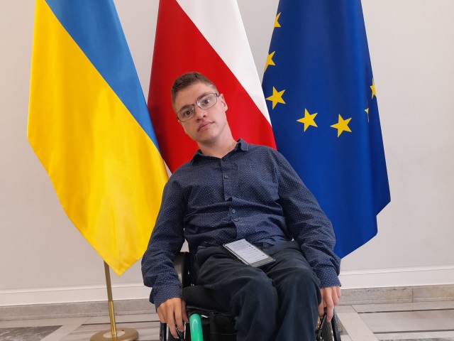 Młody mężczyzna na wózku stoi na tle flag Polski, Unii Europejskiej i Polski.