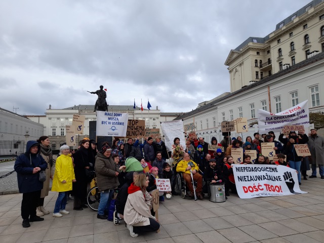 Grupowe zdjęcie uczestników protestu przed pałacem prezydenckim. Protestujący trzymają transparenty z napisami m.in. Małe "Domy Muszą być w ustawie", "Niepdagagogicnze, niezauważalne Dość tego!" , "Godnego życia dla opiekunek osób z niepełnosprawnością. Socjalny Kongres Kobiet Protest 2119 