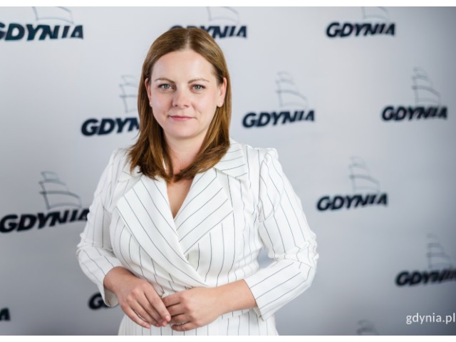 Prezdentka Gdynia, Aleksandra Kosiorek, ubrana w jasną marynarkę stoi na tle baneru z napisami Gdynia. 