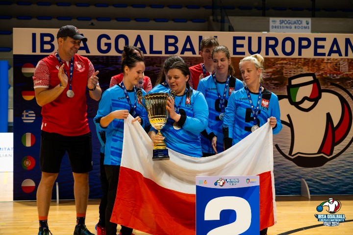 Reprezentantki Polski w goal ball. Cztery kobiety, w niebieskich bluzkach reprezentacji. Jedna z nich trzyma w ręku puchar. Pozostałe trzymają biało-czerwona flagę. 