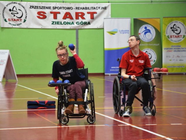 kobieta na wózku rzuca piłkę z bocci, obok patrzy na nią mężczyzna na wózku. Są na hali sportowej, podczas konkursu bocci