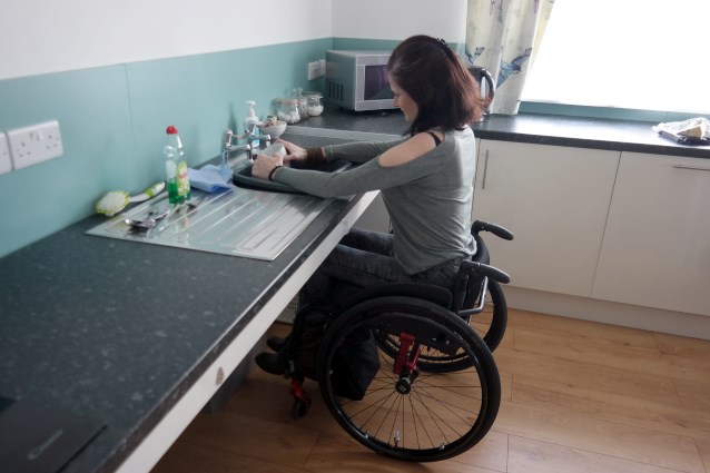 Kobieta na wózku myje kubek, przestrzeń pod blatem jest niezabudowana, co pomaga osobom poruszającym się na wózkach na bezpieczne użytkowanie kuchni