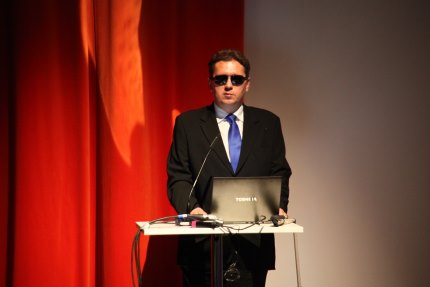 Piotr Witek, w czarnych okularach i czarnym garniturze, przemawia podczas konferencji, stojąc za pulpitem z laptopem