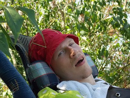 Twarz młodego mężczyny, leżącego na wózku wśród zielonych liści krzewów