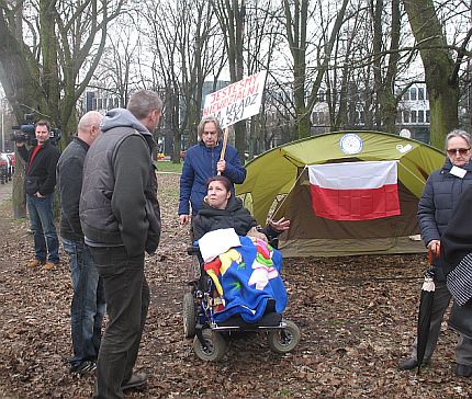 Manifestacja. Kobieta na wózku mówi do dwóch meżczyzn. W tle namiot z flagą Polski