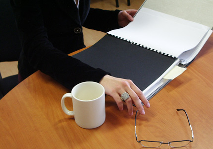 Stół w biurze, ręce kobiety w garsonce otwierają notatnik, obok stoi kubek