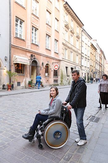 Uliczka na Starówce. Kobieta siedzi na wózku, który pcha mężczyzna