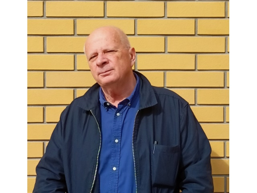 Michał Brutkowski, mężczyzna w starszym w wieku, ubrany w niebieską koszulę i kurtkę stoi na tle ściany.