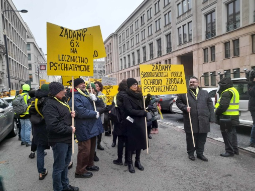 Grupa ludzi stoi na ulicy trzymając transparenty m.in. z napisami "Żądamy 6 procent na lecznictwo psychiatryczne" i "Żądamy środowiskowego modelu ochrony zdrowia psychicznego".