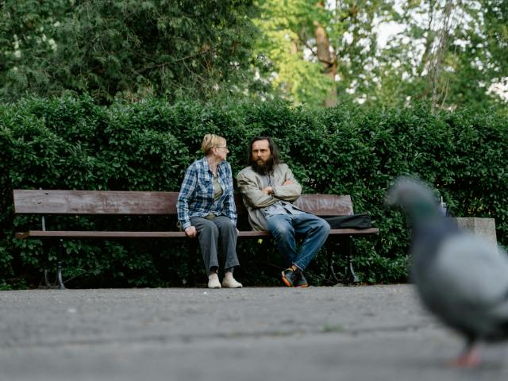 Kobieta i mężczyzna w średnim wieku, będący w kryzysie bezdomności siedzą na ławce i toczą ze sobą rozmowę.